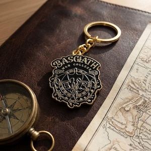 May include: A gold-toned metal keychain featuring a shield-shaped emblem with black enamel inlay. The emblem displays the text BASGIATH WAR COLLEGE and a central dragon graphic with wings spread, surrounded by intricate floral and vine-like filigree.