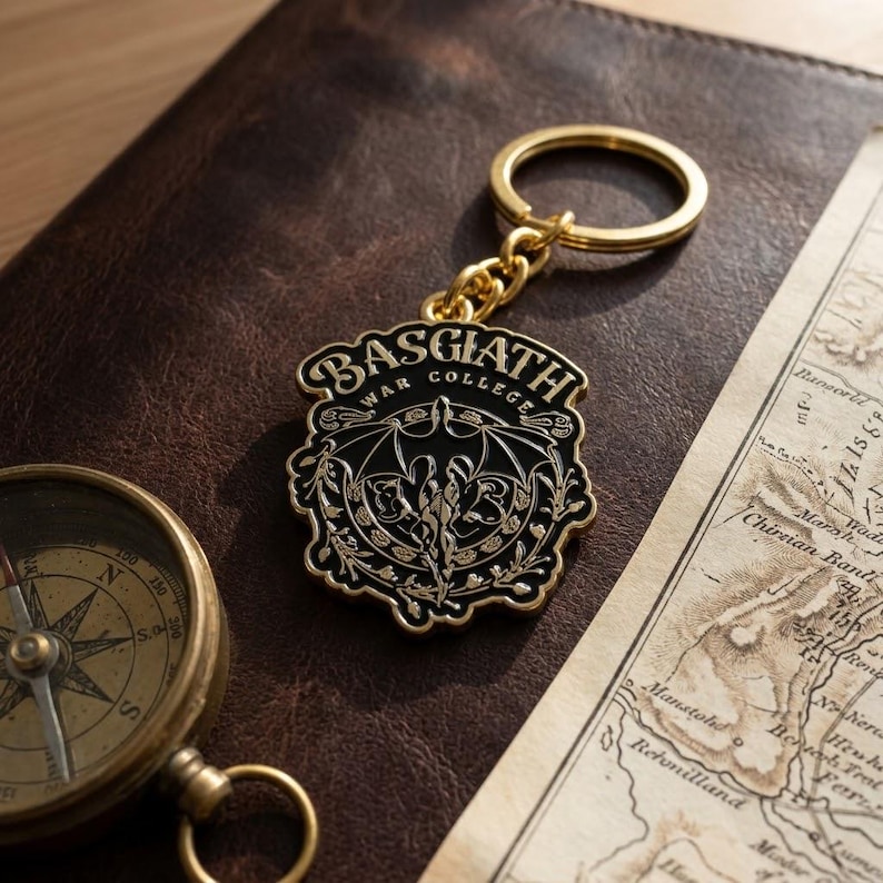 May include: A gold-toned metal keychain featuring a shield-shaped emblem with black enamel inlay. The emblem displays the text BASGIATH WAR COLLEGE and a central dragon graphic with wings spread, surrounded by intricate floral and vine-like filigree.