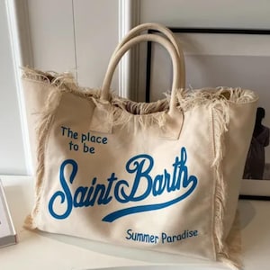 May include: A beige canvas tote bag with two top handles and frayed edges. The bag features blue text that reads The place to be, Saint Barth, and Summer Paradise. The design includes a large script font for the words Saint Barth.