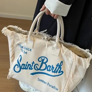 May include: A cream-colored canvas tote bag with frayed edges and two top handles. The bag features blue printed text that reads "The place to be", "Saint Barth", and "Summer Paradise" on the front. The bag has an open top design.