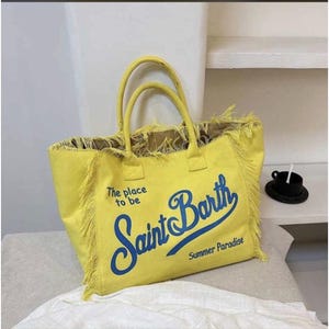Puede incluir: Un bolso tote de color amarillo brillante con dos asas y bordes deshilachados. El frente presenta el texto azul "The place to be", "Saint Barth" y "Summer Paradise" en letra cursiva. El bolso tiene una forma rectangular estructurada.