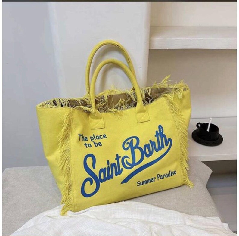 Puede incluir: Un bolso tote de color amarillo brillante con dos asas y bordes deshilachados. El frente presenta el texto azul "The place to be", "Saint Barth" y "Summer Paradise" en letra cursiva. El bolso tiene una forma rectangular estructurada.