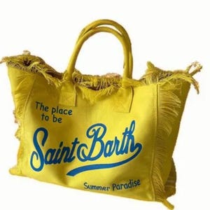 Puede incluir: Un bolso tote de lona en color amarillo brillante con dos asas superiores y bordes deshilachados. La parte frontal presenta texto azul que dice The place to be Saint Barth Summer Paradise en una mezcla de fuentes impresas y cursivas.