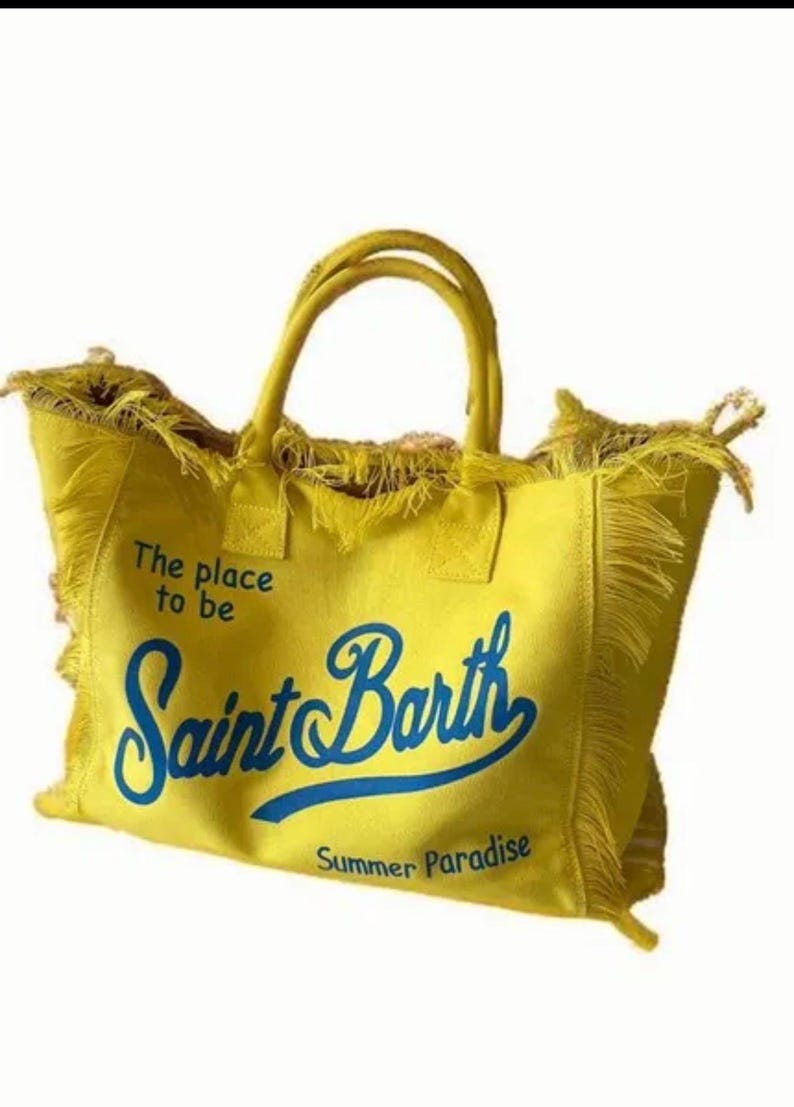 Puede incluir: Un bolso tote de lona en color amarillo brillante con dos asas superiores y bordes deshilachados. La parte frontal presenta texto azul que dice The place to be Saint Barth Summer Paradise en una mezcla de fuentes impresas y cursivas.