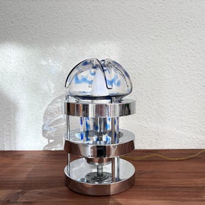 May include: A modern table lamp with a clear glass shade featuring blue accents. The lamp has a cylindrical chrome base with three circular tiers. A gold-colored cord extends from the base.