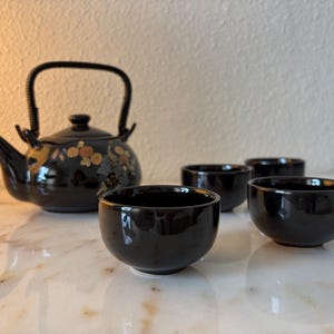Op de afbeelding: Een zwarte keramische theeset bestaande uit een theepot met een gebogen handvat en een bloemmotief in oranje, gele en blauwe tinten, vergezeld van drie bijpassende zwarte keramische theekopjes die voor de theepot zijn geplaatst.