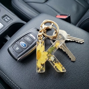 May include: A gold-coloured keyring with the letter "K" made of clear resin, embedded with yellow flowers and gold flakes. The keyring is attached to a set of keys and a car key fob.