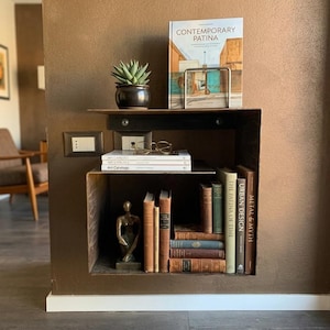 May include: A built-in, dark brown bookshelf with a variety of items. A potted succulent, a book titled "CONTEMPORARY PATINA", and a pair of glasses sit on the top shelf. The lower shelf holds books and a small bronze statue.