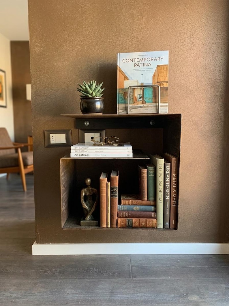 May include: A built-in, dark brown bookshelf with a variety of items. A potted succulent, a book titled "CONTEMPORARY PATINA", and a pair of glasses sit on the top shelf. The lower shelf holds books and a small bronze statue.