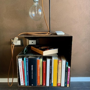 May include: A unique industrial-style bedside table with a built-in bookshelf. It features a black metal frame, a large Edison bulb lamp, and a collection of books. The lamp has a brown cord. The table is made of dark wood.
