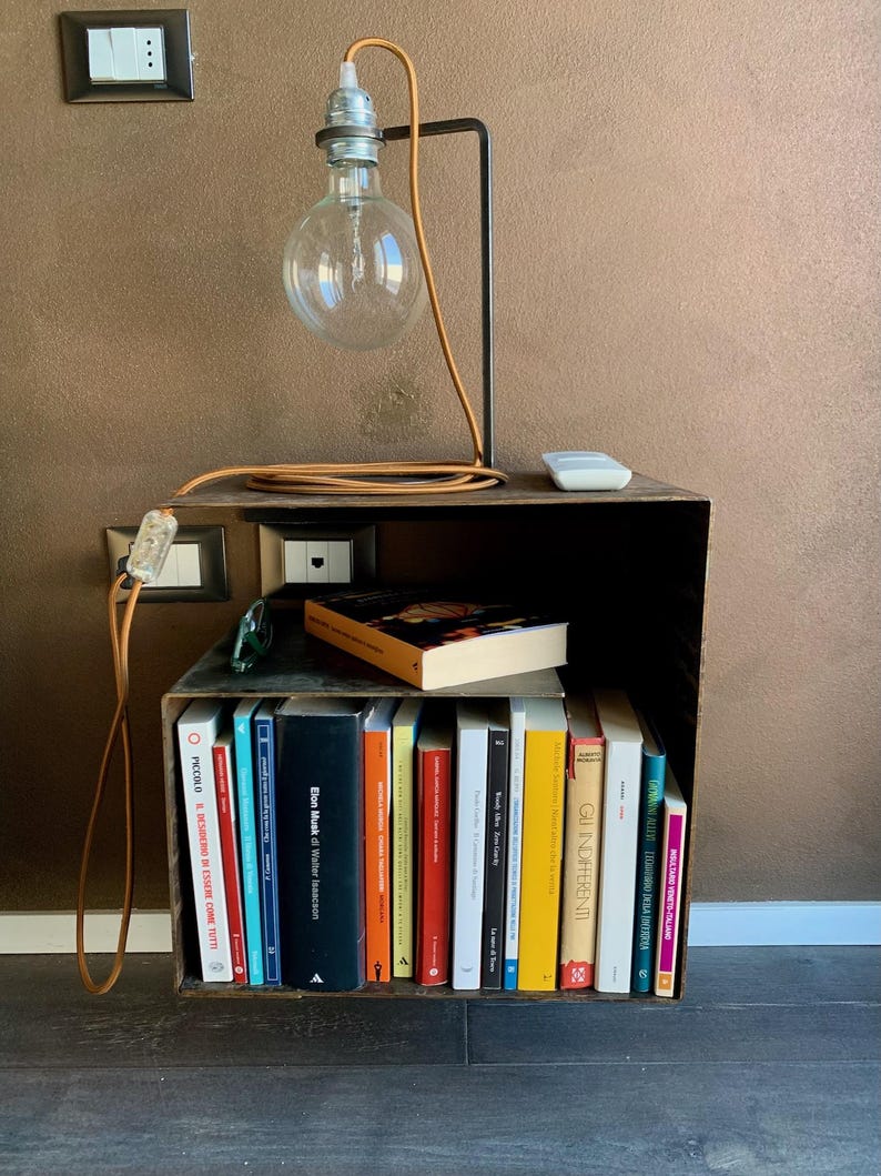 May include: A unique industrial-style bedside table with a built-in bookshelf. It features a black metal frame, a large Edison bulb lamp, and a collection of books. The lamp has a brown cord. The table is made of dark wood.