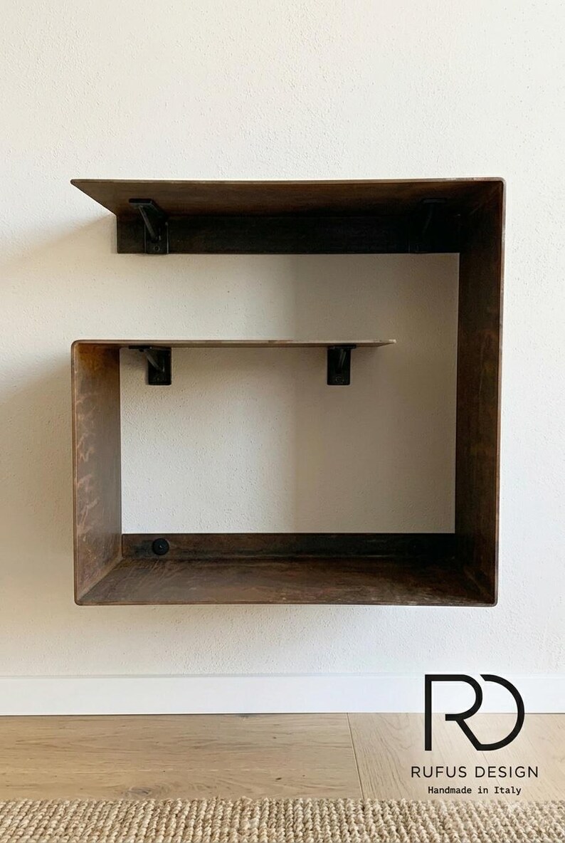 May include: A modern, wall-mounted shelf unit with a dark brown, weathered finish. The design features a large, open square frame with a smaller shelf inside, supported by black metal brackets. The bottom right corner displays the text "RD RUFUS DESIGN Handmade in Italy."