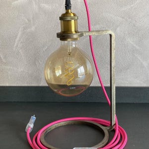 Op de afbeelding: Een unieke bureaulamp met een grote, ronde Edison-lamp. De lamp heeft een messingkleurige lamphouder en een roze elektrisch snoer. De basis is een cirkelvormig metalen frame en het snoer bevat een snoerschakelaar.