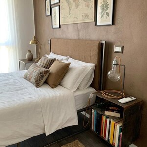 May include: Bedroom interior with a bed featuring white linens, decorative pillows, and a brown headboard. A side table with a lamp and books sits beside the bed. Artwork and a world map hang on the brown wall.