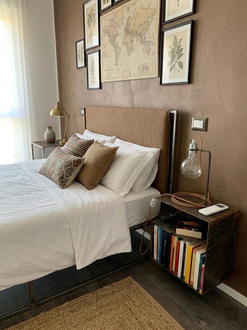 May include: Bedroom interior with a bed featuring white linens, decorative pillows, and a brown headboard. A side table with a lamp and books sits beside the bed. Artwork and a world map hang on the brown wall.