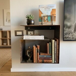 May include: A modern, industrial-style wall shelf made of dark metal. It holds a small potted succulent, books, a sculpture, and a book titled "CONTEMPORARY PATINA". The shelf is mounted on a neutral-colored wall, creating a minimalist aesthetic.