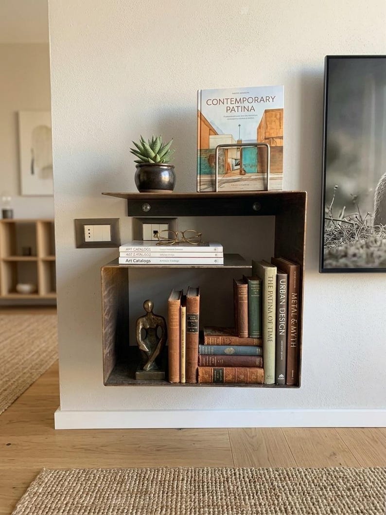May include: A modern, industrial-style wall shelf made of dark metal. It holds a small potted succulent, books, a sculpture, and a book titled "CONTEMPORARY PATINA". The shelf is mounted on a neutral-colored wall, creating a minimalist aesthetic.