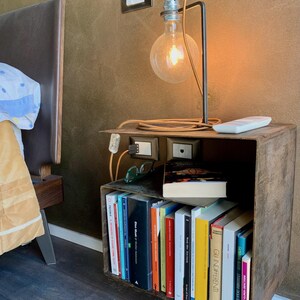 May include: A rustic bedside table made from a repurposed metal box, holding a collection of books. A modern lamp with a large, exposed bulb illuminates the scene. The lamp is connected to a power outlet on the table. A remote control and glasses are also on the table.