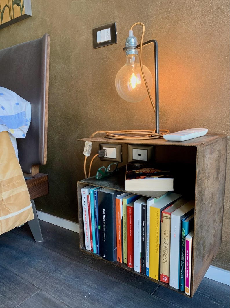 May include: A rustic bedside table made from a repurposed metal box, holding a collection of books. A modern lamp with a large, exposed bulb illuminates the scene. The lamp is connected to a power outlet on the table. A remote control and glasses are also on the table.
