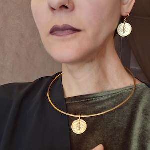 Könnte beinhalten: Goldfarbenes Schmuckset mit Halskette, Ohrringen und Armband. Jedes Teil hat einen runden Anhänger mit einem Bienenmuster. Die Halskette ist ein einfacher Choker. Die Ohrringe sind hängend, und das Armband ist ein Armreif.