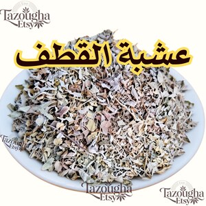 May include: A white bowl filled with dried, shredded plant leaves in shades of beige, light brown, and pale green. The Arabic text "عشبة القطف" is printed in large yellow letters above the bowl, with the brand name "Tazougha Etsy" visible.