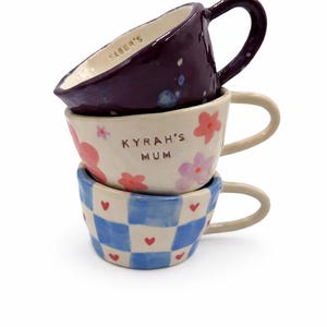 May include: A stack of three ceramic mugs. The top mug is dark purple with the text "SABER'S" inside. The middle mug is cream-coloured with pink and purple flowers and the text "KYRAH'S MUM". The bottom mug features a blue and white checkerboard pattern.