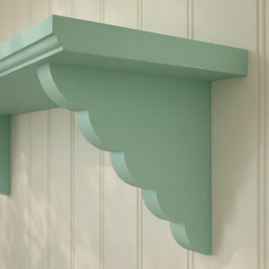 May include: A mint green wooden shelf with decorative scalloped brackets. The shelf is mounted on a white panelled wall. The shelf has a classic design and is suitable for displaying items.