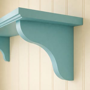 May include: A pair of light blue wooden shelves with decorative curved brackets. The shelves are mounted on a cream-coloured wall with vertical panelling. The shelves are painted in a solid, matte finish.