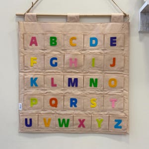 May include: A beige fabric alphabet chart with colorful letters A-Z in individual pockets. The chart is suspended by a wooden dowel and twine. The letters are in various colors, including pink, green, yellow, and blue.