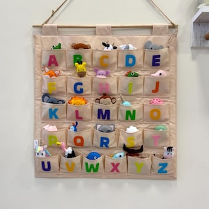 May include: A beige fabric wall organiser with pockets, each labelled with a letter of the alphabet and containing a small animal toy. The organiser hangs from a wooden dowel and a natural fibre cord. The animals are various colours.
