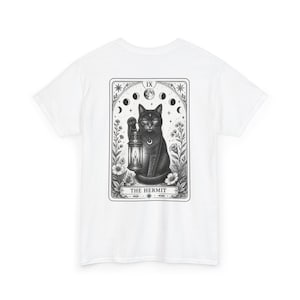 May include: A white t-shirt featuring a black and white graphic print on the back. The design depicts a cat holding a lantern within a tarot card frame labeled THE HERMIT, surrounded by floral elements and a lunar cycle phase illustration.