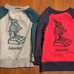 May include: Two children's sweatshirts with a microscope graphic and the word "Scientist" printed on the front. One sweatshirt is gray with teal sleeves and the other is red with navy blue sleeves.
