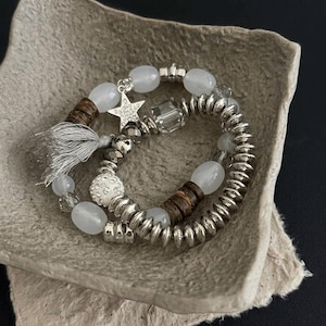 May include: Two bracelets with silver and white beads. One has a star charm and a grey tassel. The other features silver and wooden beads. The bracelets are displayed in a grey paper container.