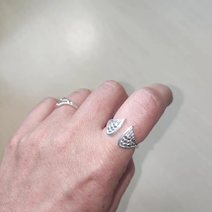 May include: A silver-colored open ring featuring two symmetrical wing-shaped ends with detailed feather-like textures. The ring is worn on the finger, positioned alongside a second, thinner, plain silver-colored band on the adjacent finger.
