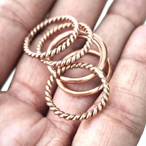Pode incluir: Um conjunto de quatro anéis cor de cobre, incluindo dois anéis com uma textura torcida semelhante a uma corda e dois anéis com um acabamento de banda lisa e simples. Os anéis são metálicos com um tom castanho-avermelhado quente.