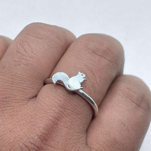 May include: A silver-toned ring featuring a small, flat squirrel silhouette as the centerpiece. The squirrel is depicted in profile with its tail curved upward. The band of the ring is thin, smooth, and polished, encircling the finger securely.