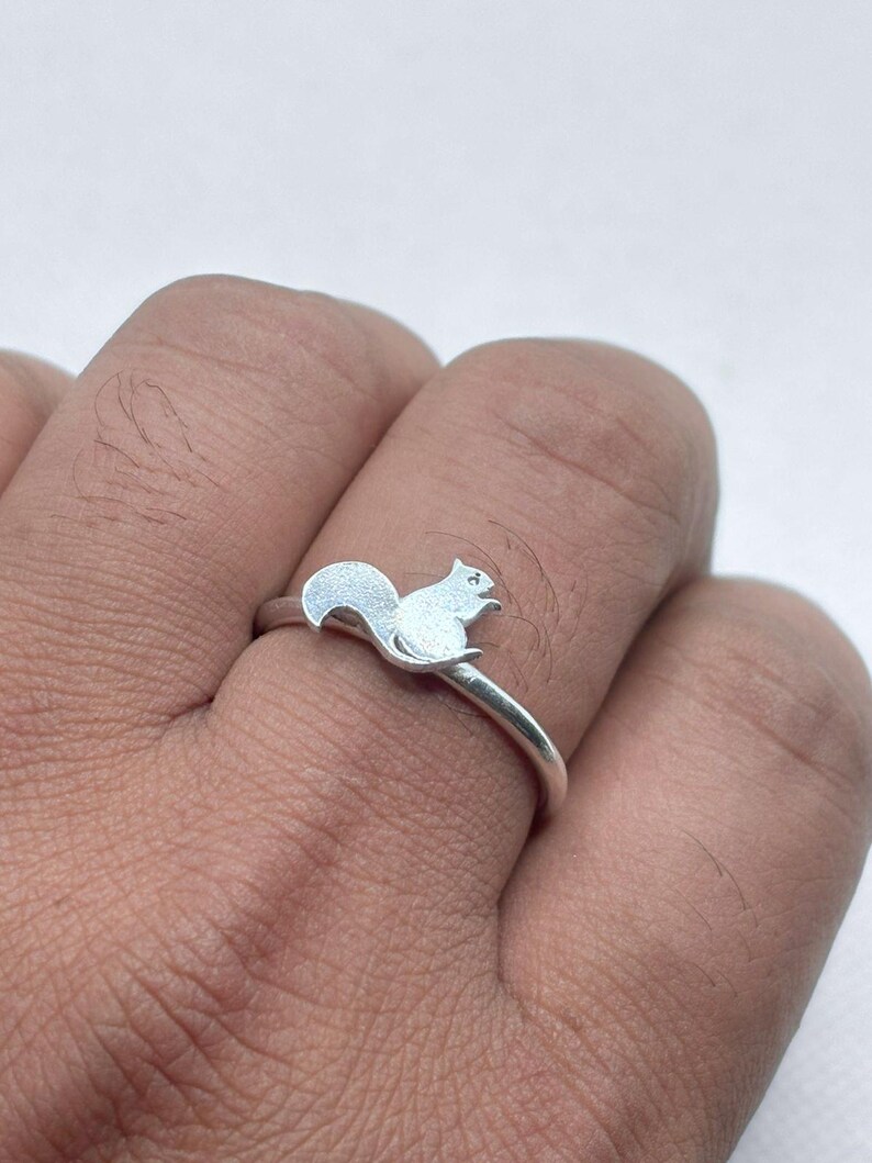 May include: A silver-toned ring featuring a small, flat squirrel silhouette as the centerpiece. The squirrel is depicted in profile with its tail curved upward. The band of the ring is thin, smooth, and polished, encircling the finger securely.