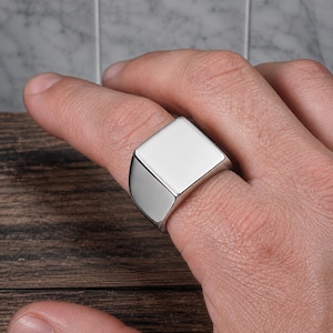 May include: A polished silver-tone signet ring featuring a smooth, flat, square-shaped face. The band is solid and tapers slightly towards the back, showcasing a clean, reflective metallic surface with no engravings or additional decorative elements.