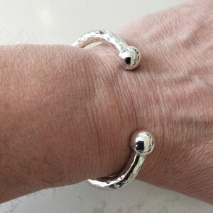 Könnte beinhalten: Ein silberfarbenes, offenes Armband mit einer texturierten, gehämmerten Oberfläche entlang des Bandes und zwei glatten, polierten kugelförmigen Enden. Das Armband ist als durchgehende Metallschleife mit einer Öffnung in der Mitte gestaltet.