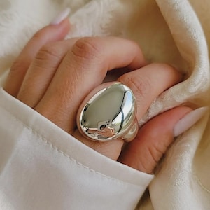 May include: A large, polished silver ring with an oval, smooth surface. The ring is worn on a finger, reflecting the surroundings. The ring's design is bold and modern, suitable for a statement piece of jewelry.