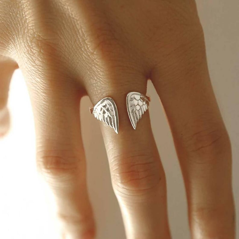 May include: A silver-toned open ring featuring two symmetrical wing-shaped ends. The metal surface displays detailed, etched feather patterns that curve inward toward the center gap, creating a delicate and polished appearance on the finger.