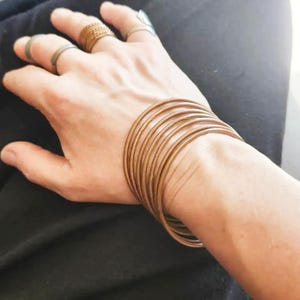 Könnte beinhalten: Ein Set aus dünnen, kupferfarbenen Metallarmreifen, die als Stapel am Handgelenk getragen werden, ergänzt durch drei passende dünne Ringe an den Fingern. Der Schmuck hat eine glatte, polierte Oberfläche und ein schlichtes, kreisförmiges Design.