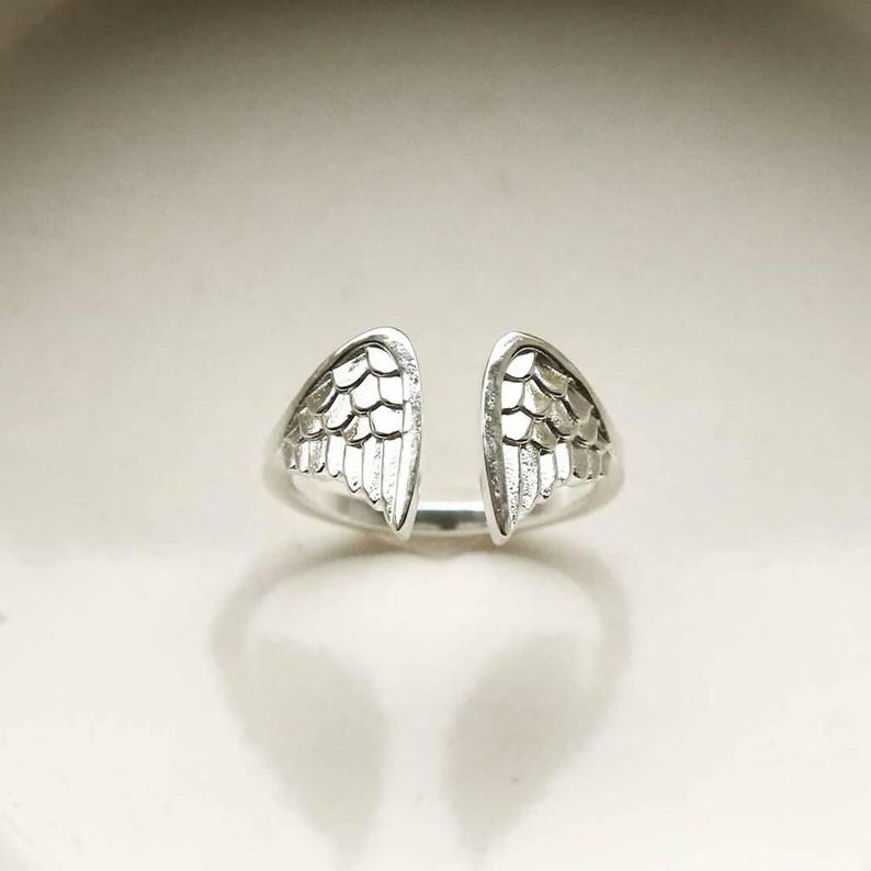 May include: A silver-toned open ring featuring two symmetrical wing-shaped motifs facing each other. The wings are detailed with engraved, overlapping feather patterns that create a textured appearance on the metallic surface of the band.