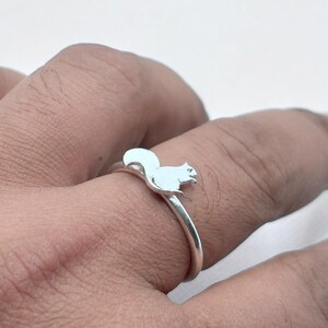 May include: A silver-toned ring featuring a small, flat squirrel silhouette attached to the top of the thin, circular band. The squirrel is depicted in a seated position with its tail curved upward, showcasing a smooth, polished metallic finish.