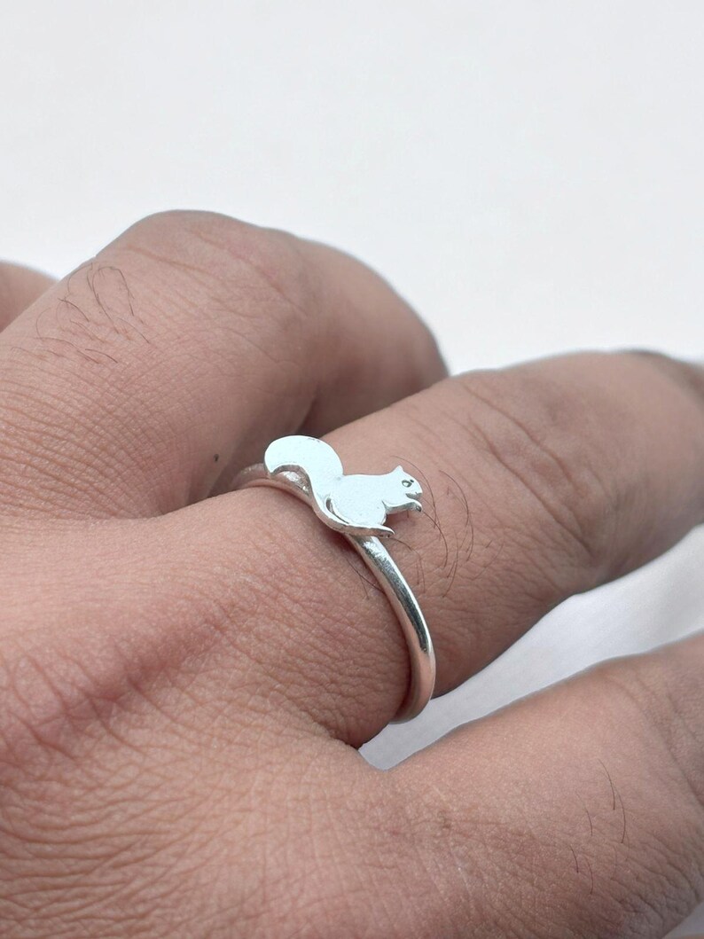 May include: A silver-toned ring featuring a small, flat squirrel silhouette attached to the top of the thin, circular band. The squirrel is depicted in a seated position with its tail curved upward, showcasing a smooth, polished metallic finish.