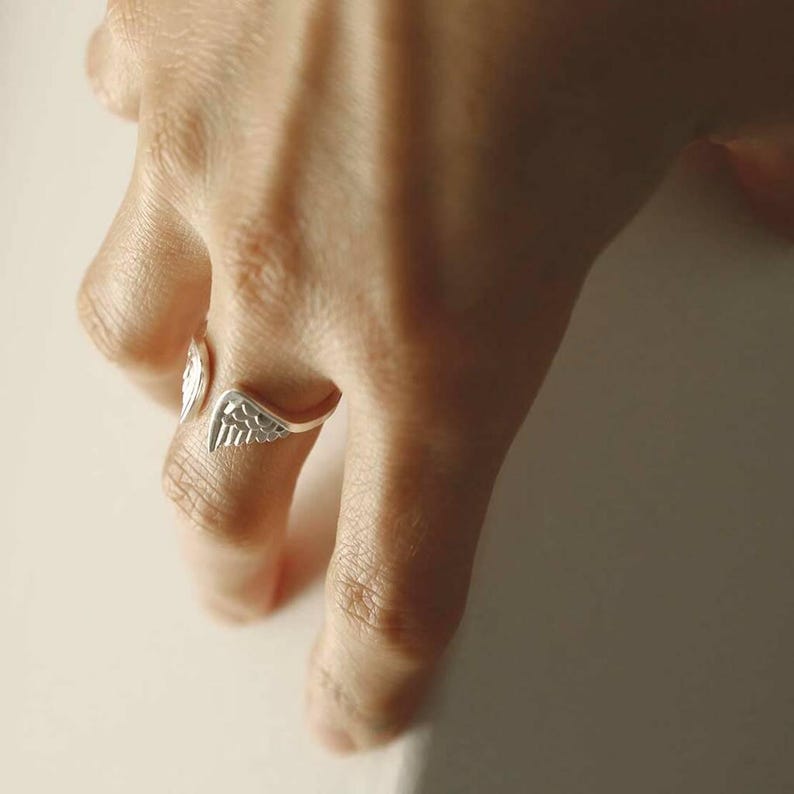 May include: A silver-toned open ring featuring two detailed wing motifs that wrap around the finger. The metal band is polished and smooth, with engraved lines on the wings to define the feathers. The ring is adjustable, showing a gap between the wings.