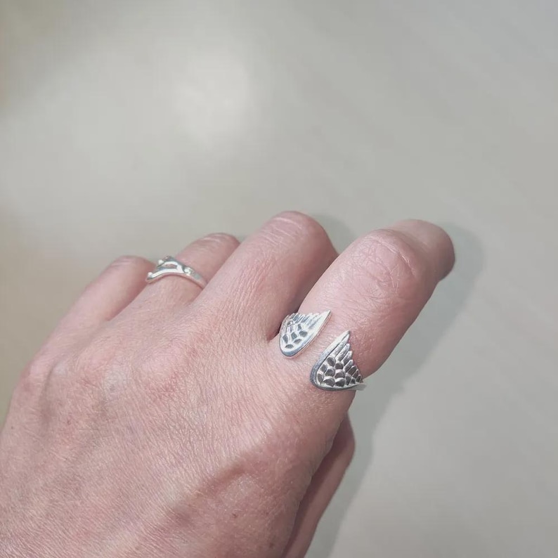 May include: A silver-colored open ring featuring two wing-shaped ends with detailed feather textures, worn on the index finger, alongside a second simple silver-colored band on the ring finger, both displaying a polished metallic finish.