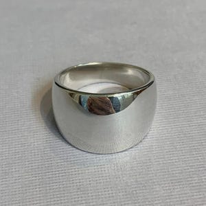 May include: A polished, silver-colored ring with a wide band. The ring has a smooth, rounded surface that reflects light. The ring is a simple, yet elegant piece of jewelry.