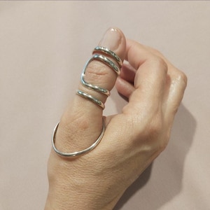 May include: A silver-colored metal splint ring designed to wrap around the thumb. The piece features a continuous, smooth wire construction that circles the base of the thumb and extends upward with two additional loops that encircle the upper joint.
