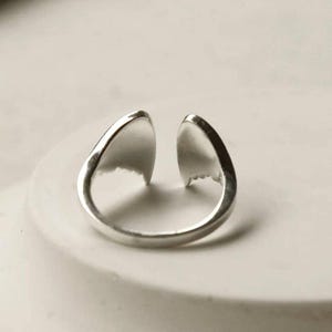 May include: A silver-toned open ring featuring two symmetrical, pointed, wing-like elements that face each other at the top. The band is smooth and polished, creating a continuous loop that remains open at the center, showcasing a metallic finish.